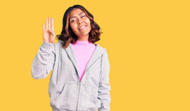 Genç, güzel, melez, sportif kıyafetler giyen, dört numaralı parmaklarıyla işaret eden, kendinden emin ve mutlu bir şekilde gülümseyen bir kadın.. 