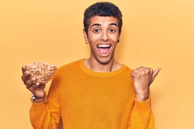 Fıstık tutan genç Afrikalı Amerikalı adam başparmağıyla yan tarafı işaret ediyor ve ağzı açık gülümsüyor. 