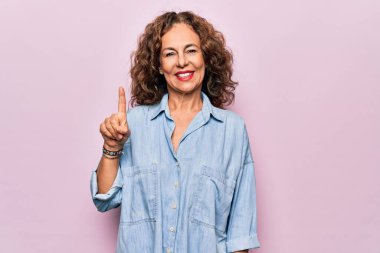 Orta yaşlı güzel bir kadın pembe arka planda dikilen sıradan bir kot gömlek giyer ve bir numaralı parmağını göstererek gülümserken kendinden emin ve mutlu olur..