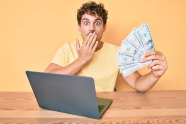 Kıvırcık saçlı, bilgisayar bilgisayarı kullanarak çalışan genç beyaz bir adam elinde ABD doları banknotları tutarak ağzını eliyle kapatıyor, şok geçiriyor ve hata yapmaktan korkuyor. şaşırmış ifade 