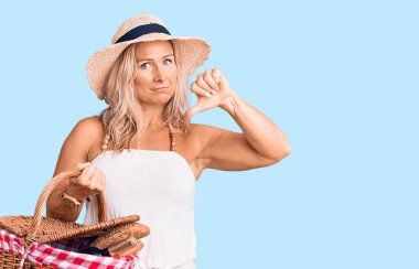 Orta yaşlı, sarışın, yaz şapkası takan ve elinde piknik sepeti olan kızgın yüzlü, olumsuz işaretli, baş parmaklı, reddedilmiş bir kadın. 