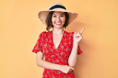 Yazlık şapka takan güzel bir kız gülümsüyor mutlu bir şekilde elini ve parmağını yana doğru gösteriyor. 