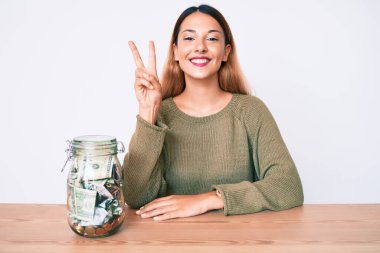 Masada içi dolar dolu bir kavanozla oturan genç esmer kadın kameraya gülümseyen ve zafer işareti yapan. İki numara.. 