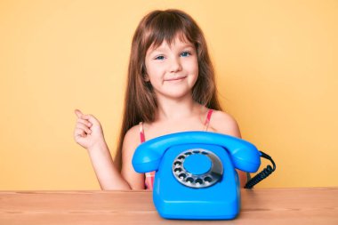 Masada oturan, uzun saçlı, klasik telefon kullanan, mutlu gülümseyen, elini ve parmağını yana doğru gösteren beyaz bir kız çocuğu. 
