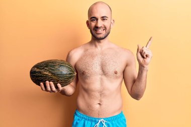 Genç, kel, mayo giyen, elinde kavun tutan, gülümseyen, mutlu, eli ve parmağı yanda olan bir adam. 