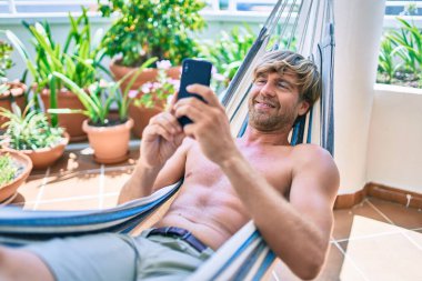 Orta yaşlı yakışıklı bir adam evinin terasında, bir hamakta telefonla dinleniyor.