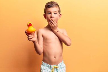 Mayo giyen sevimli sarışın çocuk elinde ördek oyuncağı, ağzını eliyle kapatıyor, şokta ve hata yapmaktan korkuyor. şaşırmış ifade 