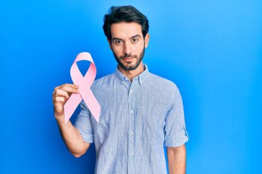 Young hispanic man holding pink cancer ribbon thinking attitude and sober expression looking self confident  clipart