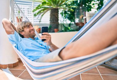 Orta yaşlı yakışıklı bir adam evinin terasında, bir hamakta telefonla dinleniyor.