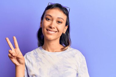 Gündelik kıyafetler giyen, gülümseyen ve kameraya göz kırpan genç bir kadın zafer işareti yapıyor. İki numara.. 