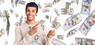 Genç Afrikalı Amerikalı sıradan kıyafetler giyen, gülümseyen ve kameraya bakıp iki elini ve parmağını yana doğru gösteren bir adam..