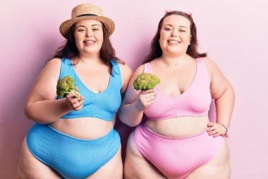 Bikini giyen ve elinde brokoli tutan genç ve büyük beden ikizler. Pozitif ve mutlu görünüyorlar. Dişlerini gösteren bir gülümsemeyle ayakta duruyorlar. 