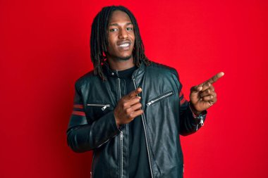 Siyah deri ceketli Afrikalı Amerikalı adam gülümsüyor ve kameraya bakıyor iki eliyle ve parmaklarıyla kenarı işaret ediyor.. 