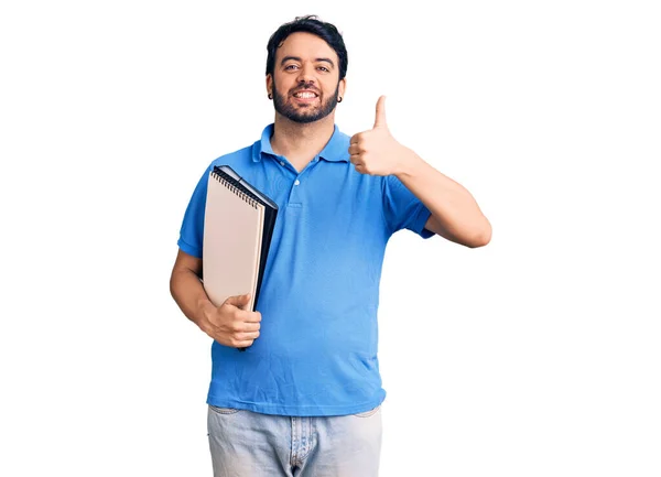 Joven Hombre Hispano Sosteniendo Cuaderno Sonriendo Feliz Positivo Pulgar Hacia —  Fotos de Stock