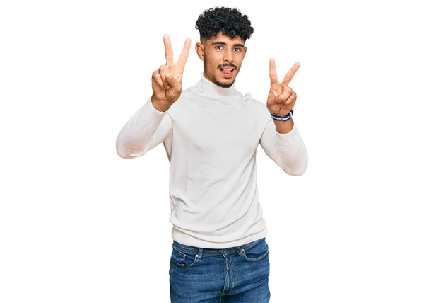 年轻的阿拉伯男子穿着休闲地冬季毛衣 伸出舌头微笑着 伸出双手的手指做胜利的标志 第二点 — 图库照片