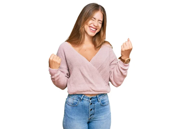カジュアルな冬のピンクのセーターを着て美しいブロンドの女性は非常に満足し 腕を上げて勝者のジェスチャーを行う興奮し 笑顔と成功のために叫んでいます お祝いのコンセプト — ストック写真