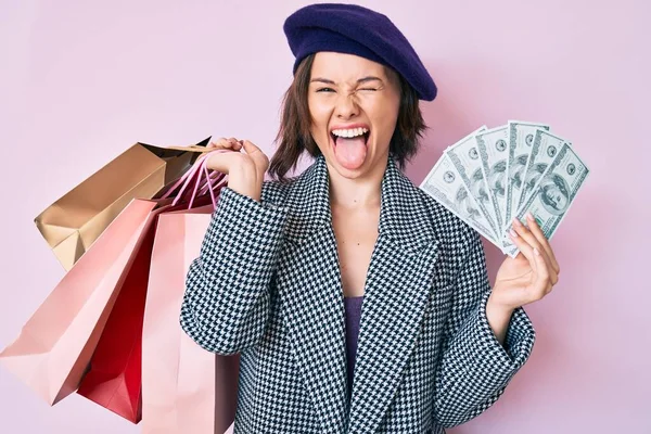 年轻美丽的女人头戴贝雷帽 提着购物袋和钞票 带着滑稽的表情伸出舌头 高兴极了 — 图库照片