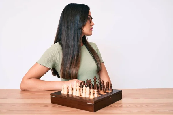 Young caucasian woman sitting at the desk playing chess looking to side, relax profile pose with natural face with confident smile.