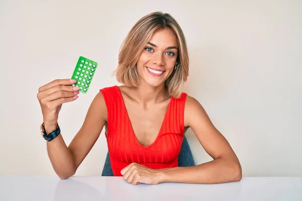 Beautiful caucasian woman holding birth control pills looking positive and happy standing and smiling with a confident smile showing teeth