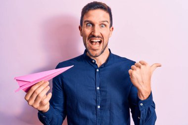 Pembe kağıt uçak tutan genç yakışıklı adam izole bir arka planda duruyor ve başparmağıyla yan tarafa bakıyor mutlu bir şekilde gülümsüyor.