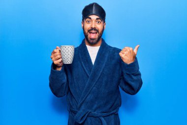 Uyku maskesi takmış, sabahlık giymiş, kahvesini yukarıyı işaret eden, ağzı açık gülümseyen genç İspanyol bir adam. 