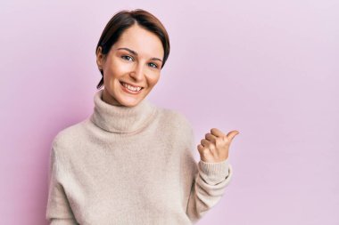 Kısa saçlı, kısa saçlı, kışlık süveter giyen neşeli yüzüyle gülümseyen ve baş parmağıyla yüzünü gösteren genç esmer kadın.. 