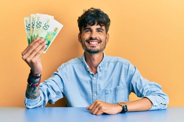 Masada oturan İspanyol asıllı genç adam pozitif ve mutlu görünüyor. Kendine güvenen bir gülümsemeyle dişlerini gösteriyor. 