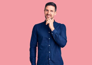 Sıradan gömlekler giyen, kameraya kendinden emin, kolları çapraz ve elleri çenesinde gülümseyen genç bir adam. Olumlu düşünmek. 