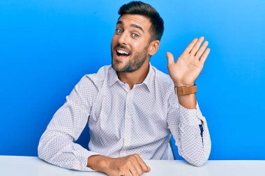 Yakışıklı İspanyol iş kıyafetleri giymiş. Masada oturmuş, mutlu, güler yüzlü, dostça bir jest yapıyor. 