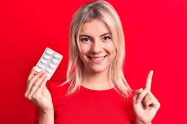 Sarışın, güzel bir kadın, izole edilmiş kırmızı arka planda durmuş haplarla gülümsüyor. Bir numara, mutlu bir yüzü işaret eden bir fikir ya da soruyla gülümsüyor.