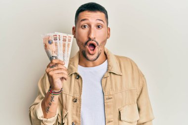 Dövmeleri olan yakışıklı bir adam. Elinde 10 pound banknotlar var. Korkmuş ve şaşırmış bir şekilde ağzı açık, inançsız bir yüz. 