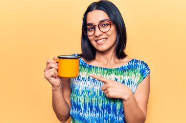 Genç, güzel, Latin bir kadın, elinde gözlük, elinde kahve, gülümsüyor. 