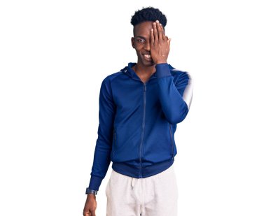 Genç Afrikalı Amerikalı erkek spor giysisi giyiyor. Bir gözünü eliyle kapatıyor. Yüzünde kendine güvenen bir gülümseme ve şaşırtıcı bir duygu var.. 