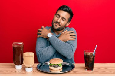 Yakışıklı İspanyol bir adam lezzetli bir klasik burger ve soda yiyor mutlu ve pozitif, kendinden emin gülümsüyor. Sevgi ve özgüven. 