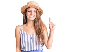 Genç, sarışın, yaz şapkası takan ve gülümseyen, kendinden emin ve mutlu bir şekilde bir numaralı parmağını gösteren bir kadın.. 