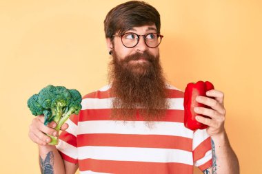 Uzun sakallı, elinde brokoli ve kırmızı biber olan yakışıklı kızıl saçlı bir adam.. 