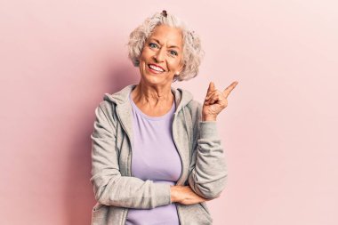 Yaşlı, gri saçlı, günlük spor kıyafetleri giyen, gülümseyen, el ve parmak uçlarını gösteren bir kadın. 