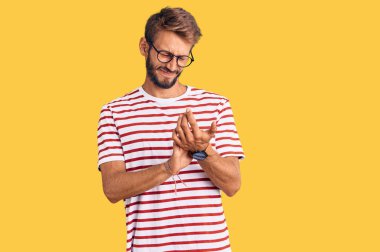 Sakallı, sıradan kıyafetler giyen, gözlüklü, elleri ve parmakları ağrıyan yakışıklı sarışın bir adam. 