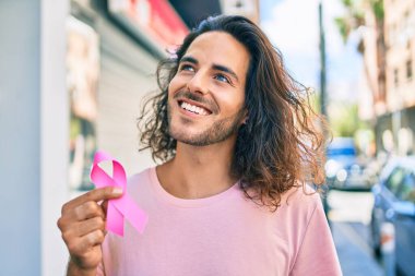 Genç İspanyol gülümseyen adam şehirde göğüs kanseri pembe kurdele tutuyor..