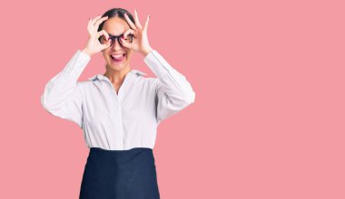 Güzel, esmer, profesyonel garson önlüğü giyen genç bir kadın dürbün gibi jest yapıyor. Dilini dışarı çıkarıyor, gözleri parmaklarından bakıyor. çılgın ifade. 