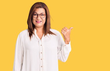 Orta yaşlı, güzel bir kadın gözlük takıyor ve parmaklarıyla yüzünü işaret ediyor.