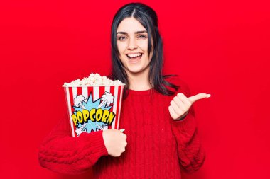 Patlamış mısır yiyen genç ve güzel bir kız başparmağıyla yan tarafa bakıyor. Ağzını açıp gülümsüyor. 