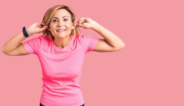 Spor kıyafetleri giymiş genç sarışın kadın gülümsüyor kulaklarını parmaklarıyla çekiyor, komik bir jest. deneme sorunu 