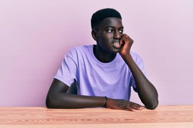 Gündelik giysiler giymiş genç Afro-Amerikan bir adam masada oturmuş gergin ve gergin görünüyor, elleri ağızdan tırnak yiyor. anksiyete. 