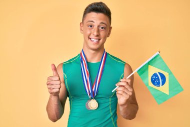 Spor kıyafetleri giyen, Brezilya bayrağı taşıyan mutlu ve pozitif gülümseyen, mükemmel ve onay işareti yapan İspanyol asıllı genç. 