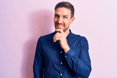 İzole edilmiş pembe arka planda duran rahat gömlekli genç bir adam kameraya kollarını kavuşturup çenesine dokunarak gülümsüyor. Olumlu düşünüyorum..