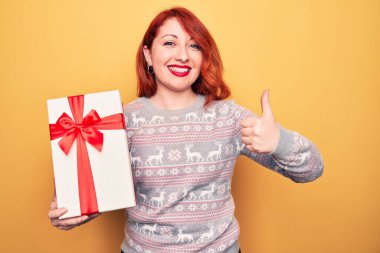 Genç, güzel, kızıl saçlı bir kadın, izole edilmiş sarı arka planda Noel hediyesini tutuyor mutlu ve pozitif gülümsüyor, başparmağıyla mükemmel ve onay işareti yapıyor.