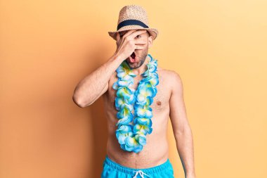 Yakışıklı, kel, mayosuz bir adam ve Hawaii Lei 'si şokla yüzünü ve gözlerini kapayarak korkuyla parmaklarının arasından bakıyor. 