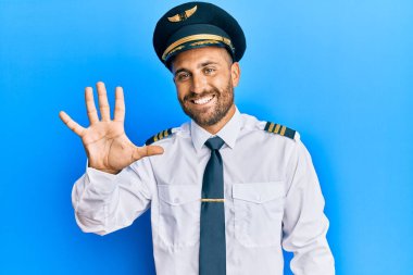 Sakallı, uçak pilotu üniforması giyen yakışıklı adam beş numarayı gösterip işaret ederken kendinden emin ve mutlu bir şekilde gülümsüyor.. 