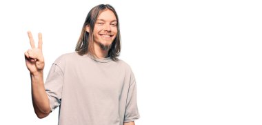 Uzun saçlı, yakışıklı beyaz bir adam. Günlük giysiler giyiyor. İki numarayı gösteriyor ve gülümsüyor.. 